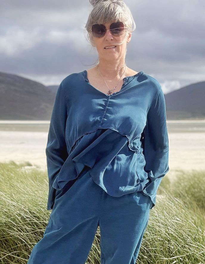 A woman wearing a matching blue satin-like top and trousers set stands outdoors in tall grass with hills and a cloudy sky in the background.