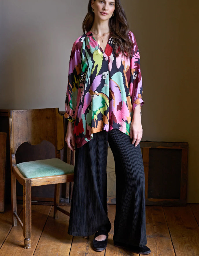 Woman wearing a colorful patterned top and black pants in a room with wooden floor and chair.