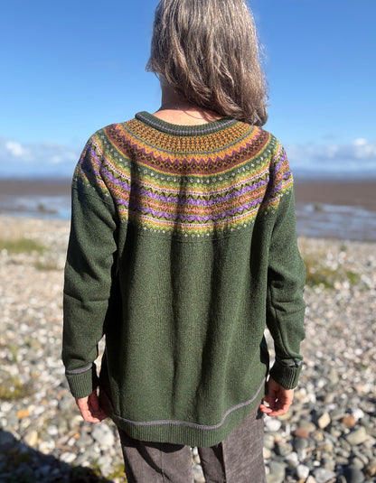 A person standing on a rocky beach, viewed from behind, wearing an olive green knit sweater with a colorful Fair Isle yoke pattern in orange, yellow, purple, and brown.