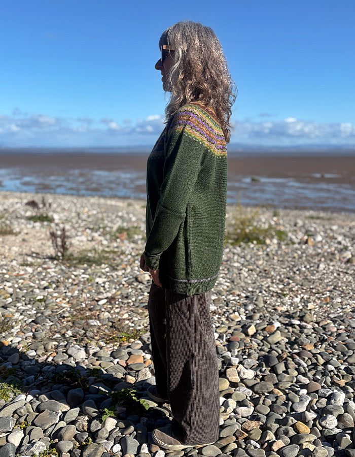 A woman stands in profile on a pebble beach wearing a dark green knit sweater with a multicolor patterned yoke and dark brown wide-leg trousers, with a blue sky and water in the background.