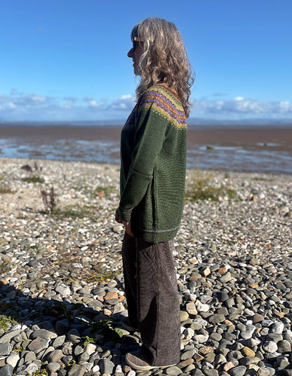 A woman stands in profile on a pebble beach wearing a dark green knit sweater with a multicolor patterned yoke and dark brown wide-leg trousers, with a blue sky and water in the background.
