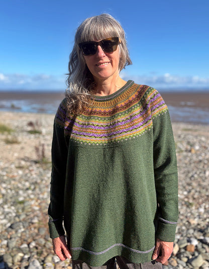 A woman wearing a green knit sweater with a colorful patterned yoke stands on a rocky beach with water and blue sky in the background.