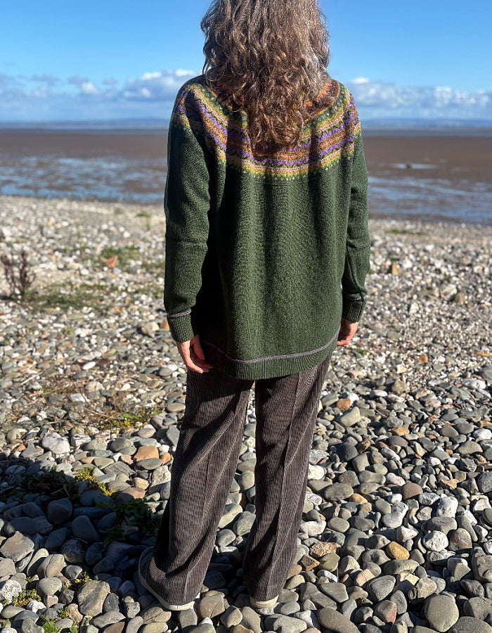 A person standing on a rocky beach, viewed from behind, wearing a dark green knit sweater with a multicolor patterned yoke and dark brown wide-leg pants.