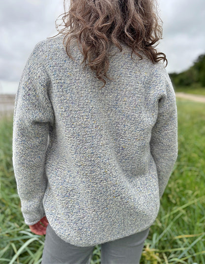 Person wearing a silver grey donegal tweed sweater and sunglasses standing in a grassy field with a cloudy sky.