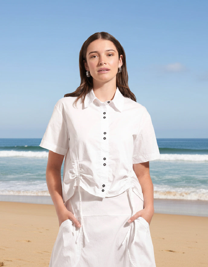 Woman in a white outfit standing on a beach with blue sky and ocean.