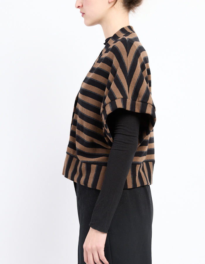 A model shown in profile wearing a cropped knit poncho-style cardigan with brown and black chevron stripes over a black long-sleeve top and black skirt, against a plain white background.