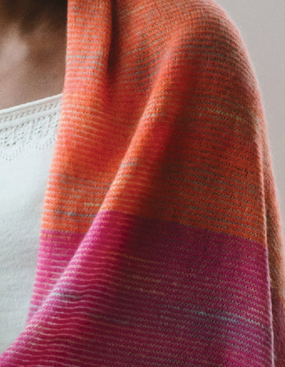 A close-up of a knitted shawl draped over a person's shoulder, featuring a color-blocked pattern with orange, pink, and magenta stripes.