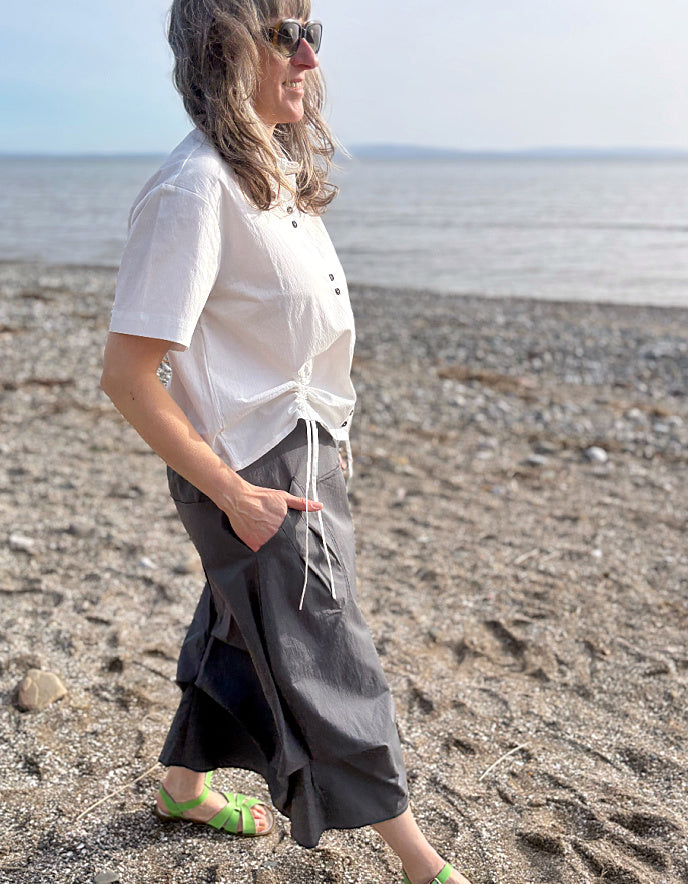 Person wearing a white shirt and gray skirt standing on a pebbly beach.