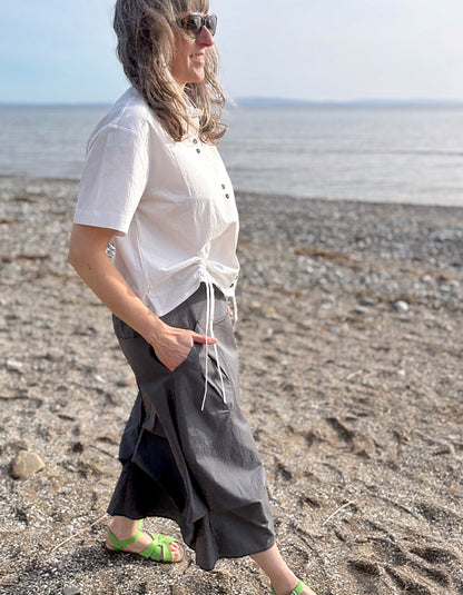 Person wearing a white shirt and gray skirt standing on a pebbly beach.