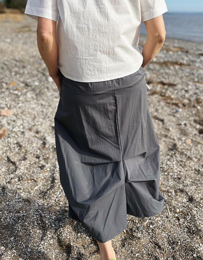 Person wearing a white shirt and gray skirt standing on a pebbly beach.