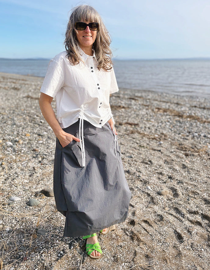 Woman standing on a beach wearing a white shirt and gray skirt with sunglasses.