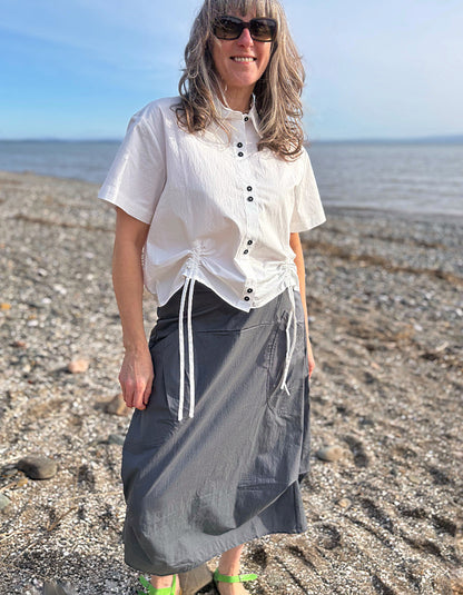 Woman standing on a beach wearing a white shirt and gray skirt with sunglasses.