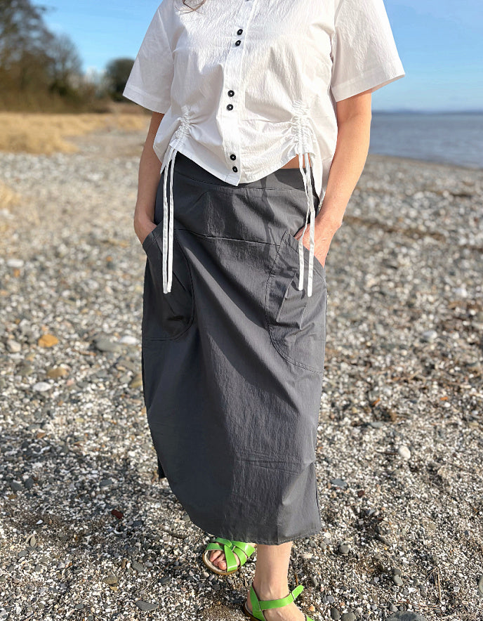 Person wearing a white shirt and gray skirt standing on a pebbly beach.