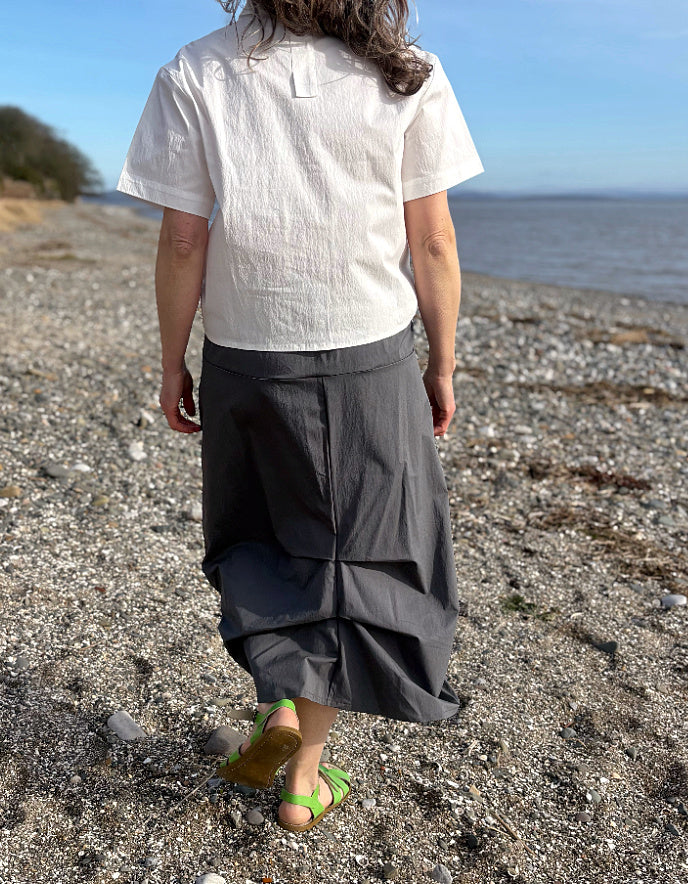Person wearing a white shirt and gray skirt walking on a pebbly beach with ocean in the background