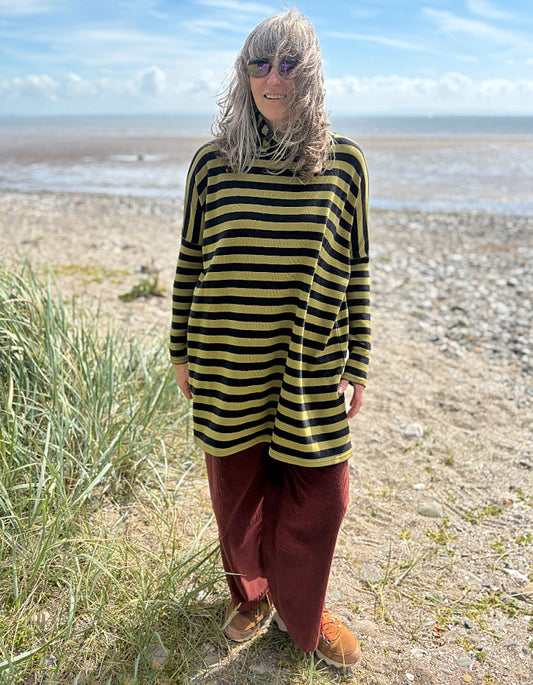 Person wearing a black and olive green striped tunic sweater and rust corduroy pants standing on a beach.