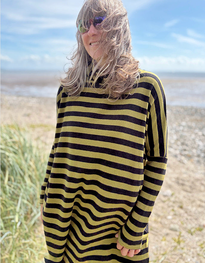 Person wearing a black and olive green striped tunic sweater and rust corduroy pants standing on a beach.