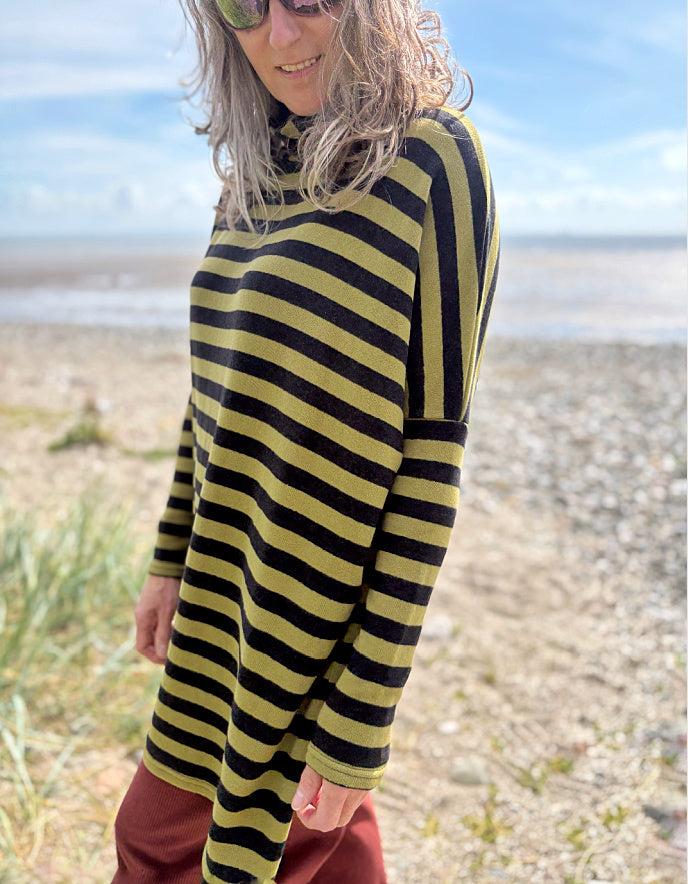 Person wearing a black and olive green striped tunic sweater and rust corduroy pants standing on a beach.