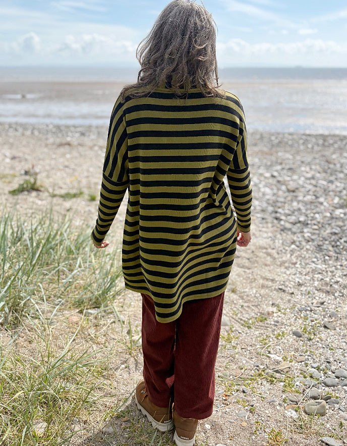 Person wearing a black and olive green striped tunic sweater and rust corduroy pants standing on a beach.