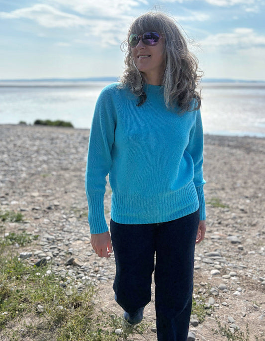 Person wearing a wool blue turquoise sweater standing in tall grass by a beach.