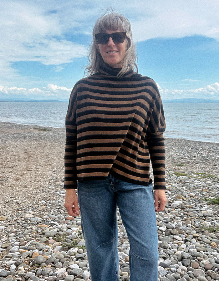 A woman with gray hair and sunglasses stands on a pebble beach wearing a brown and black horizontally striped turtleneck sweater and blue jeans, with the sea and blue sky in the background.