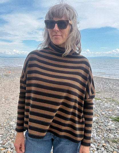 A woman with gray hair and sunglasses wearing a brown and black horizontally striped turtleneck sweater, standing on a pebble beach with the sea and sky in the background.