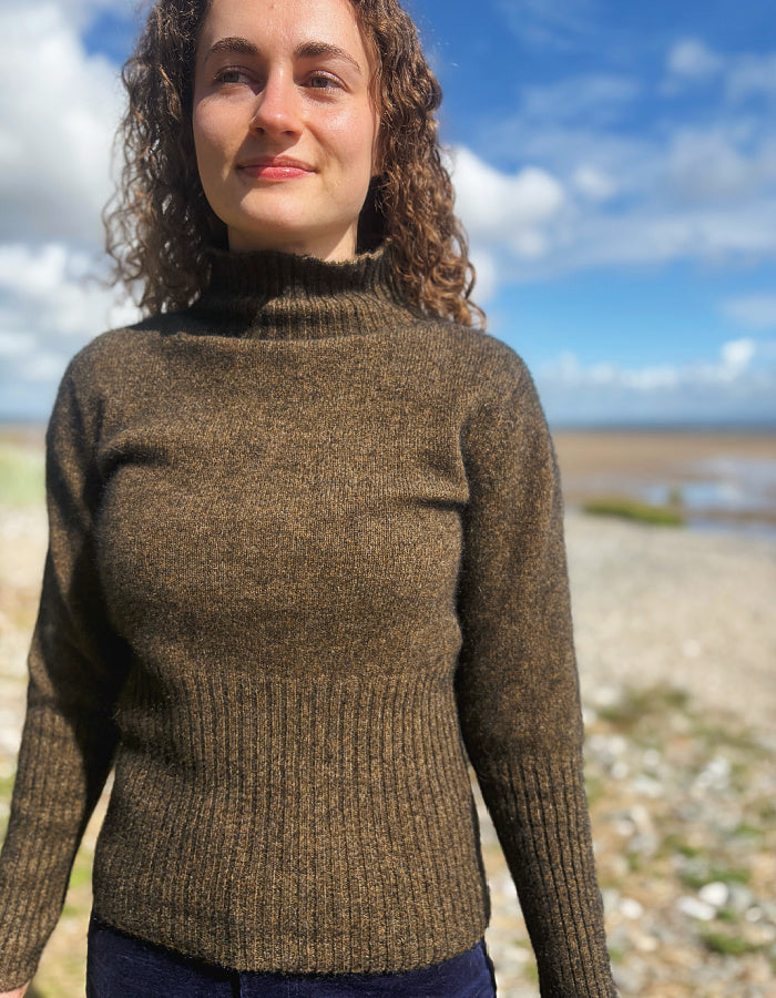 A woman with curly brown hair wearing a brown ribbed turtleneck sweater, standing outdoors on a beach with a blue sky and sea in the background.
