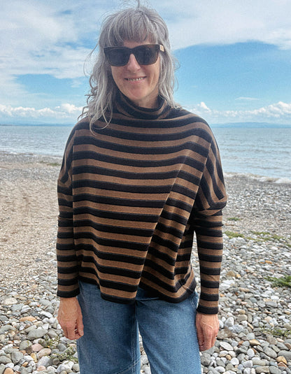 A woman with gray hair and sunglasses wearing a brown and black striped turtleneck sweater, standing on a pebble beach with the sea and blue sky in the background.