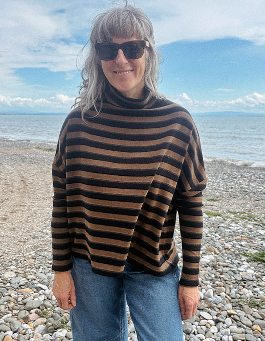 A woman with gray hair and sunglasses wearing a brown and black striped turtleneck sweater, standing on a pebble beach with the sea and blue sky in the background.