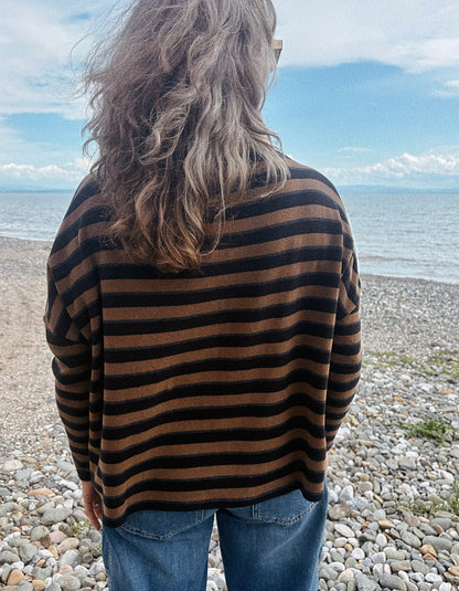 A person viewed from behind wearing a brown and black horizontally striped knit sweater and blue jeans, standing on a pebble beach with the sea and sky in the background.