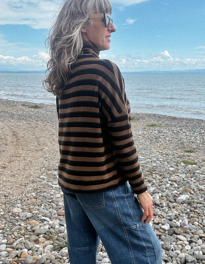 A woman with blonde hair and sunglasses stands on a pebble beach wearing a brown and black striped turtleneck sweater and blue jeans, with the sea and blue sky in the background.