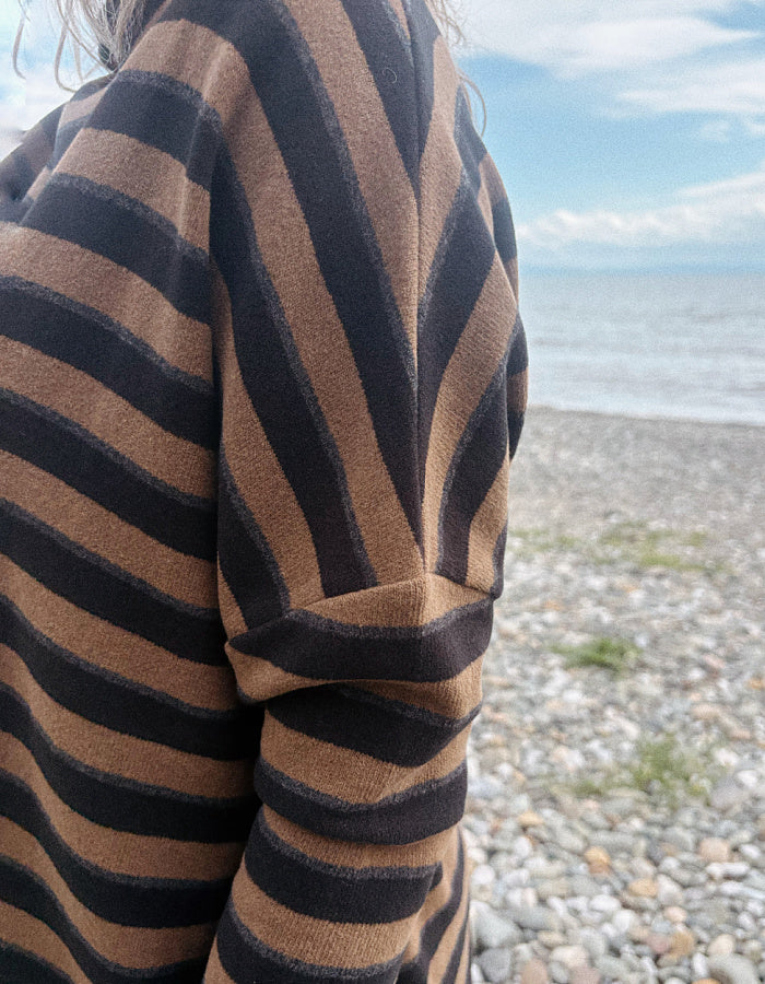 A close-up of a person wearing a brown and black striped knit sweater, standing on a pebble beach with the sea and sky in the background.
