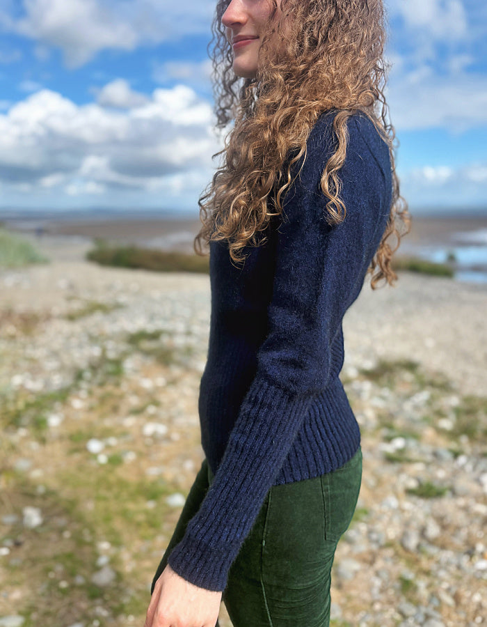 A woman with long curly light brown hair is shown from the side, wearing a dark navy blue ribbed knit sweater and dark green corduroy pants, standing outdoors on a rocky beach with a cloudy blue sky in the background.