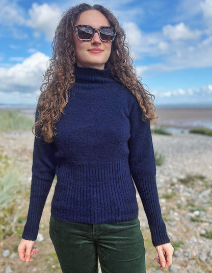 A woman with long curly brown hair wearing a navy blue ribbed turtleneck sweater and dark green pants, standing outdoors on a pebble beach with a blue sky and clouds in the background.