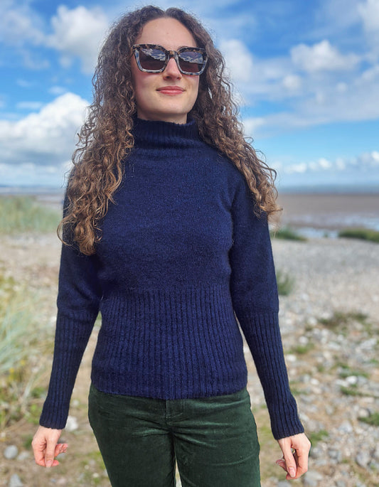 A woman with long curly brown hair wearing a navy blue ribbed turtleneck sweater and dark green pants, standing outdoors on a pebble beach with a blue sky and clouds in the background.