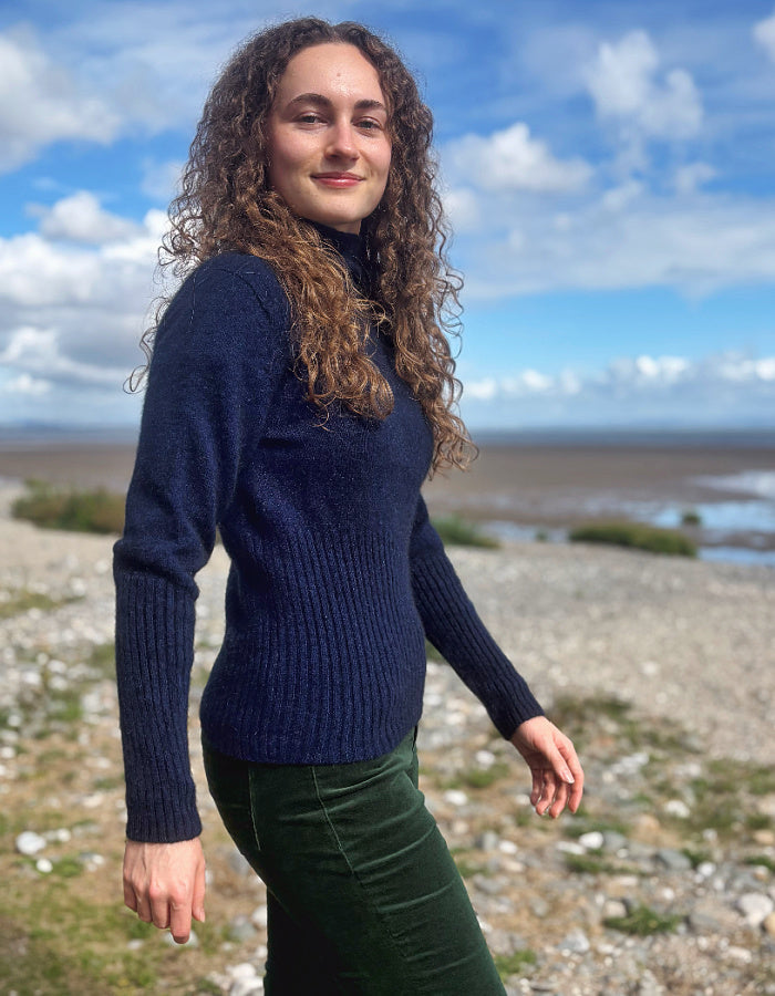 A woman with long curly brown hair stands outdoors on a rocky shoreline under a blue sky with clouds, wearing a dark navy ribbed knit sweater and dark green pants.