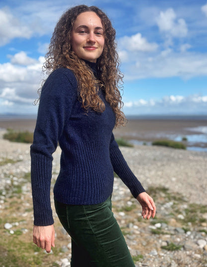 A woman with long curly brown hair stands outdoors on a rocky shoreline under a blue sky with clouds, wearing a dark navy ribbed knit sweater and dark green pants.