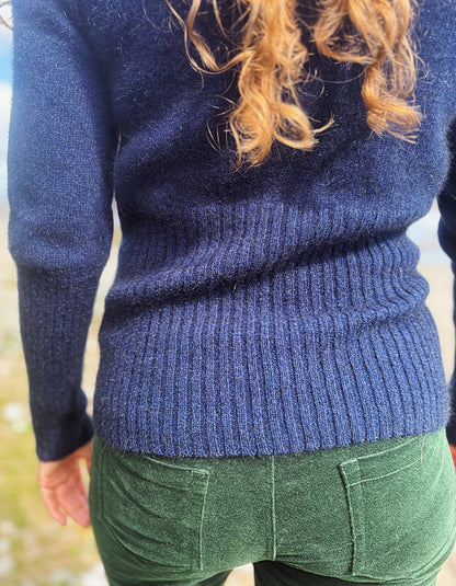 Close-up back view of a person wearing a navy blue ribbed knit sweater and green pants, with wavy light brown hair visible.