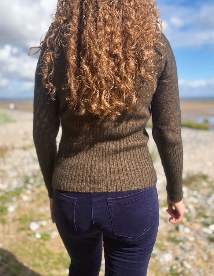 A person viewed from behind wearing a ribbed knit sweater in a dark brownish-green color, paired with dark navy pants, standing outdoors on a rocky beach with blue sky and water in the background.