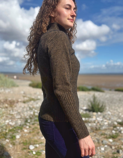 A woman with long curly brown hair stands outdoors on a pebble beach wearing a dark brown ribbed turtleneck sweater and dark blue jeans, with a cloudy blue sky in the background.
