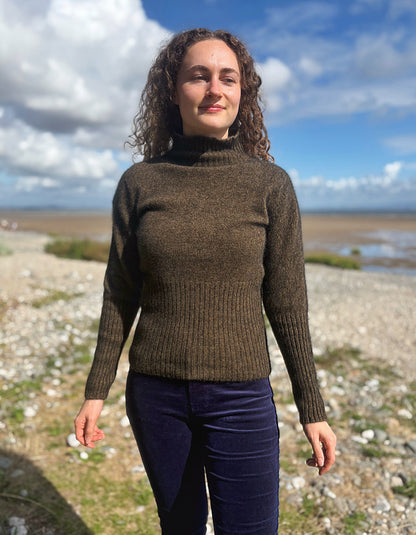 A woman with curly brown hair stands outdoors on a rocky shoreline wearing a ribbed turtleneck sweater in a dark olive-brown color and dark navy pants, with a cloudy blue sky in the background.