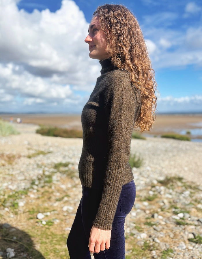 A woman with long curly brown hair stands in profile outdoors on a rocky beach, wearing a dark brown ribbed turtleneck sweater and dark navy pants under a blue sky with clouds.