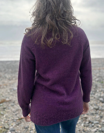 Person wearing a purple sweater standing on a pebbly beach with a cloudy sky.
