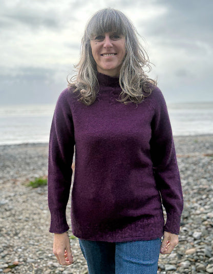 Woman wearing a purple sweater standing on a pebbly beach with water and sky in the background