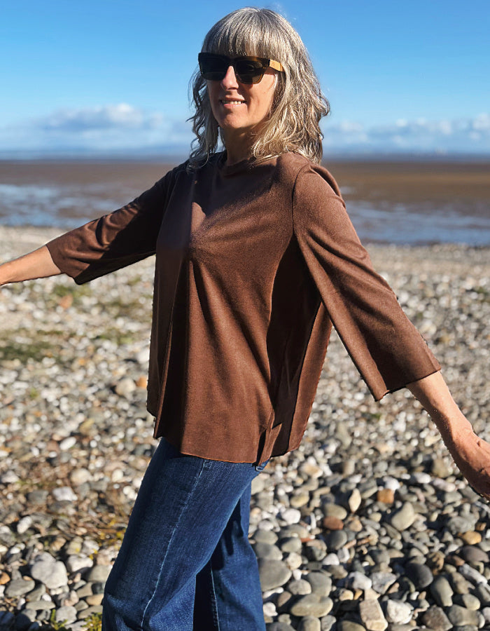A woman wearing a brown three-quarter sleeve top and blue jeans stands on a pebble beach with water and sky in the background.