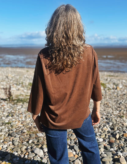 A woman with long, wavy light brown hair stands on a pebble beach, viewed from behind, wearing a brown long-sleeve top and blue jeans, with a body of water and blue sky in the background.