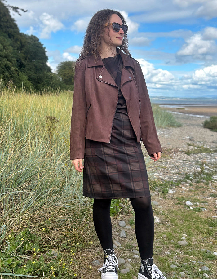 A woman stands outdoors on a grassy shoreline wearing a brown moto-style jacket, a dark plaid skirt, a black top, black tights, and white sneakers. The background shows tall grass, a pebble beach, and a cloudy blue sky.