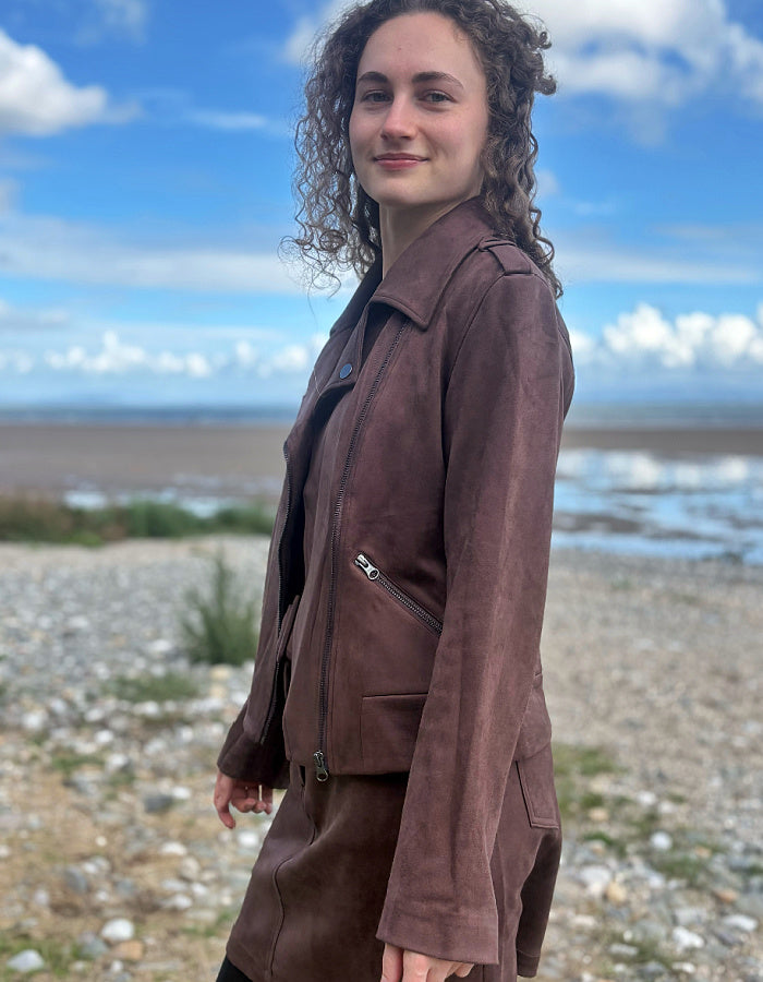 A woman with curly brown hair stands on a pebble beach wearing a brown suede jacket and matching skirt against a background of blue sky and water.
