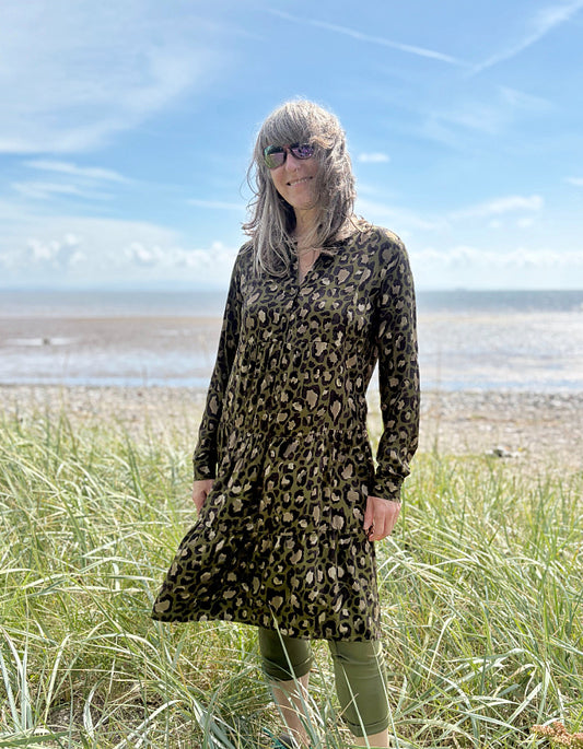 Person wearing a leopard print dress standing on a grassy field with a beach and blue sky in the background