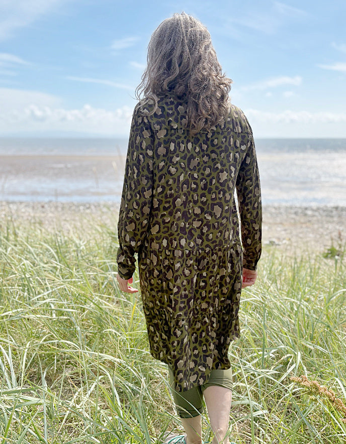 Person wearing a leopard print dress standing on a grassy field with a beach and blue sky in the background