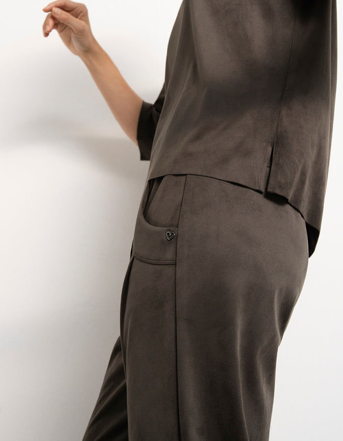 A close-up side view of a person wearing a dark olive two-piece vegan suede outfit with a short-sleeve top and matching wide-leg trousers, featuring a side pocket with a button closure.